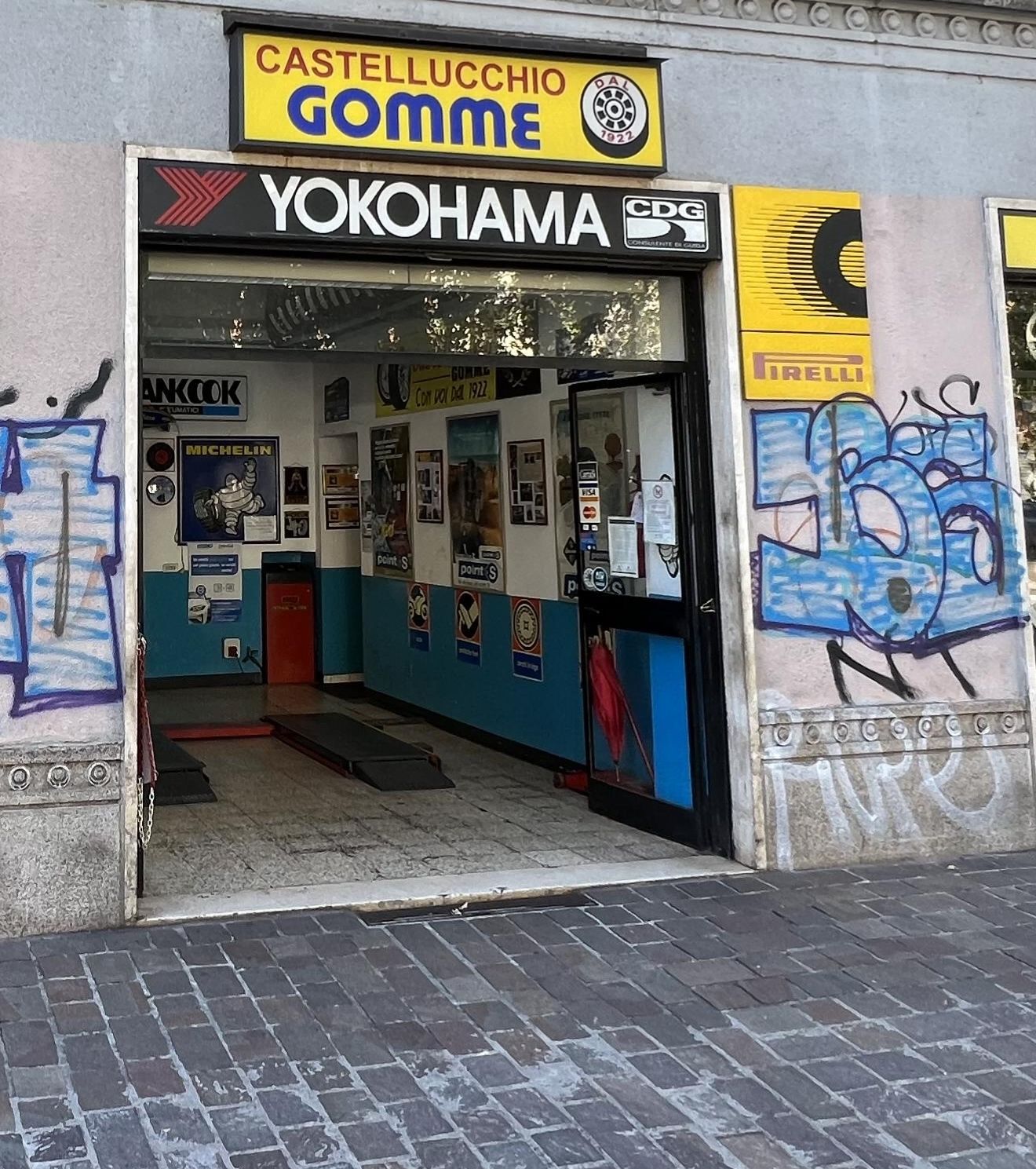 L'ingresso di IL GOMMISTA in Via Passeroni 1 a Milano: l'insegna gialla «Castellucchio Gomme dal 1922», il bandone Yokohama e la campata aperta dell'officina