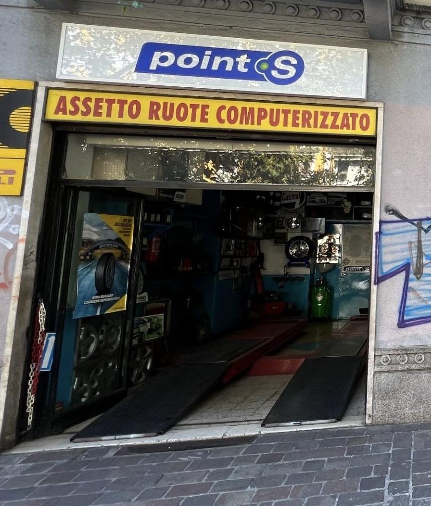 L'ingresso di Via Passeroni con il banner rosso su giallo «Assetto ruote computerizzato» sopra la campata aperta e i cerchi in vetrina
